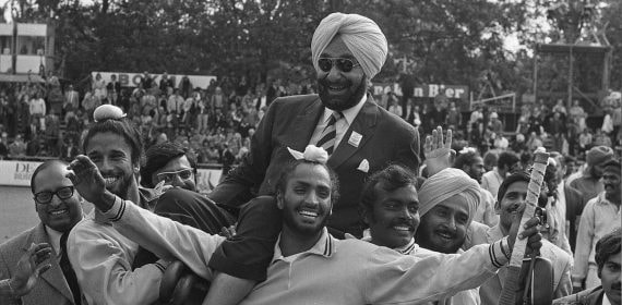 Fotografía histórica en blanco y negro de jugadores indios de hockey llevando a hombros a un directivo con turbante entre el público