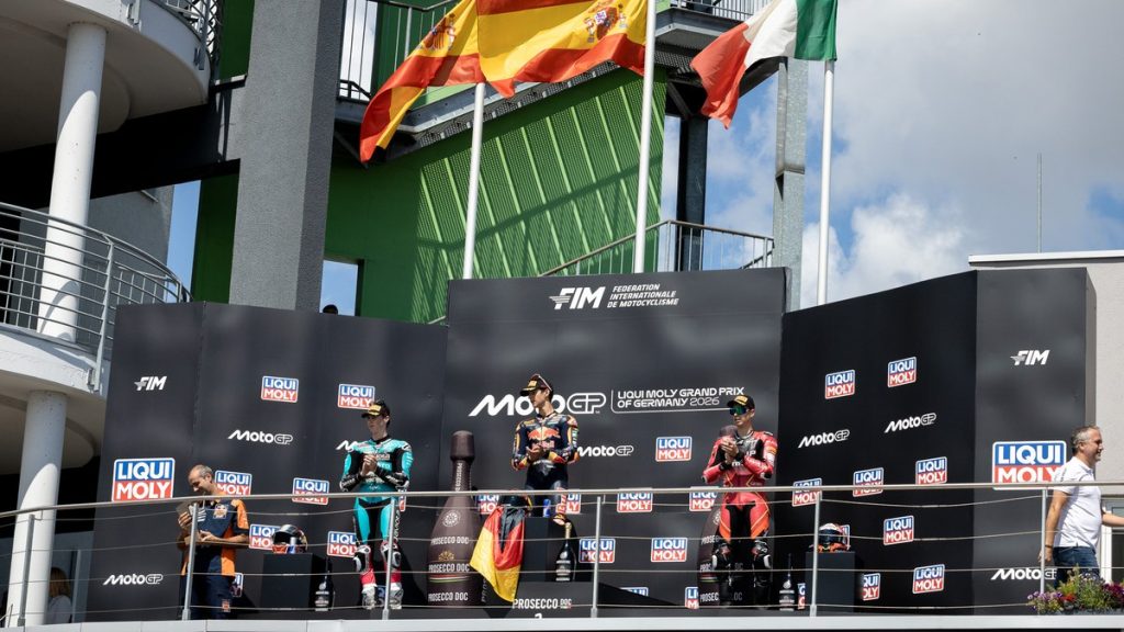 Los tres pilotos del podio de Moto3 sobre el cajón del Gran Premio de Alemania, con la bandera española ondeando detrás