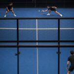 Partido de pádel visto desde detrás del cristal del fondo, con las dos parejas en pista y el público al fondo