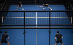 Partido de pádel visto desde detrás del cristal del fondo, con las dos parejas en pista y el público al fondo