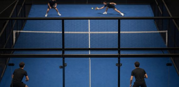 Partido de pádel visto desde detrás del cristal del fondo, con las dos parejas en pista y el público al fondo