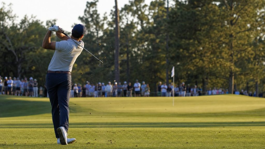 Golfista de espaldas completando el swing tras un golpe de hierro hacia el green con público en la cuerda