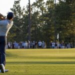 Golfista de espaldas completando el swing tras un golpe de hierro hacia el green con público en la cuerda