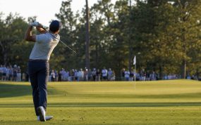Golfista de espaldas completando el swing tras un golpe de hierro hacia el green con público en la cuerda