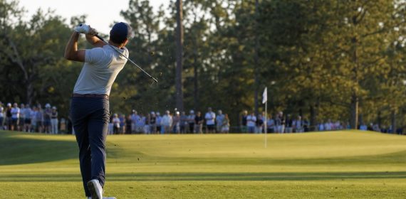 Golfista de espaldas completando el swing tras un golpe de hierro hacia el green con público en la cuerda