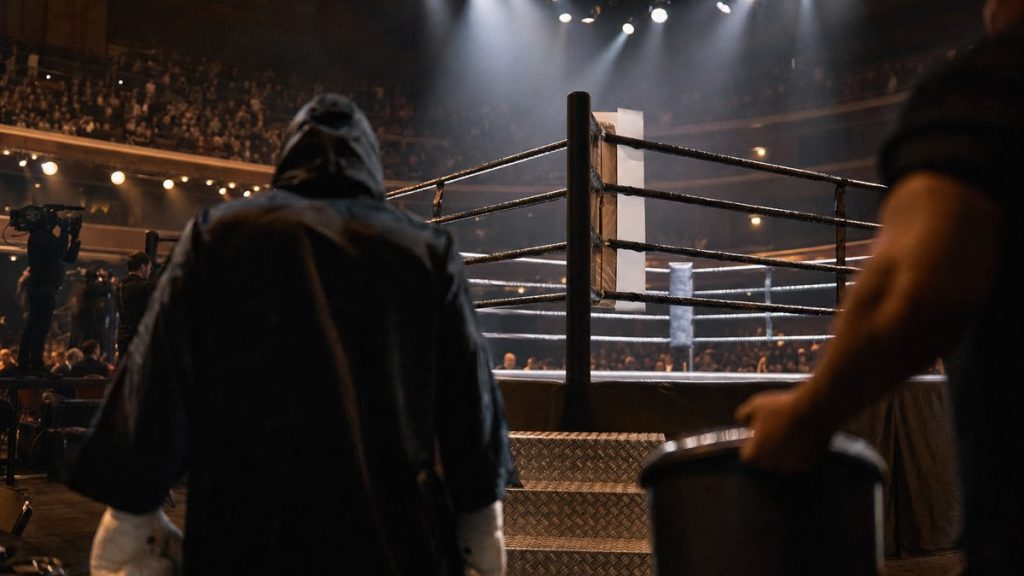 Un púgil con capucha camina hacia el ring iluminado de un pabellón antes de una velada