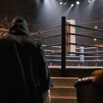Un púgil con capucha camina hacia el ring iluminado de un pabellón antes de una velada