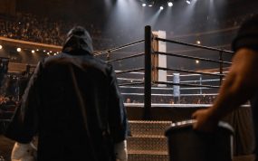 Un púgil con capucha camina hacia el ring iluminado de un pabellón antes de una velada