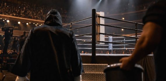 Un púgil con capucha camina hacia el ring iluminado de un pabellón antes de una velada