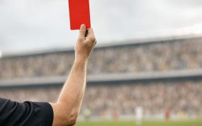El brazo de un árbitro levanta una tarjeta roja sobre el césped, con la grada desenfocada al fondo