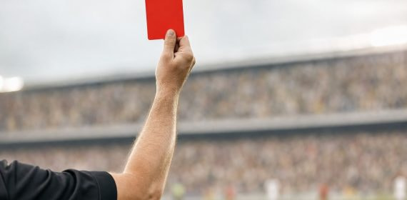 El brazo de un árbitro levanta una tarjeta roja sobre el césped, con la grada desenfocada al fondo