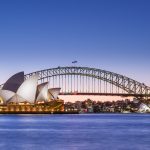 La Ópera de Sídney iluminada al anochecer, con el arco del puente de la bahía detrás y el agua del puerto en primer plano