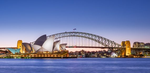 La Ópera de Sídney iluminada al anochecer, con el arco del puente de la bahía detrás y el agua del puerto en primer plano