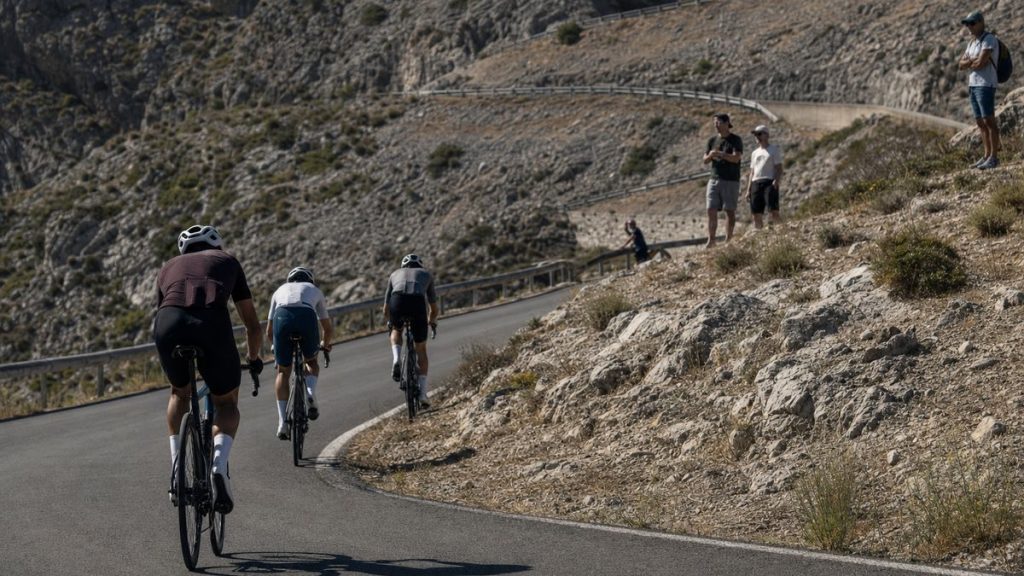 Tres ciclistas ascienden una curva de herradura en un puerto de montaña seco con público en la cuneta