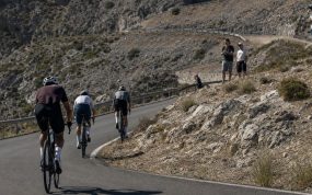 Tres ciclistas ascienden una curva de herradura en un puerto de montaña seco con público en la cuneta