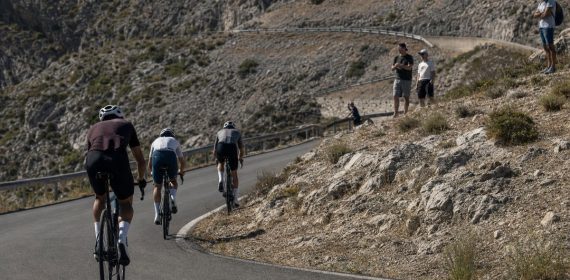 Tres ciclistas ascienden una curva de herradura en un puerto de montaña seco con público en la cuneta