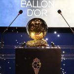 El trofeo dorado del Balón de Oro sobre un atril transparente entre dos micrófonos y un fondo azul con luces blancas