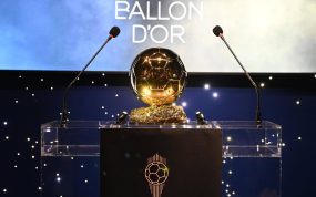 El trofeo dorado del Balón de Oro sobre un atril transparente entre dos micrófonos y un fondo azul con luces blancas