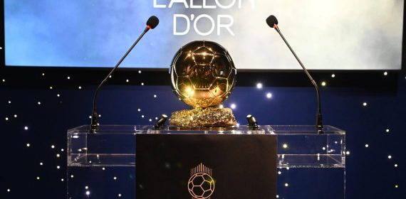 El trofeo dorado del Balón de Oro sobre un atril transparente entre dos micrófonos y un fondo azul con luces blancas