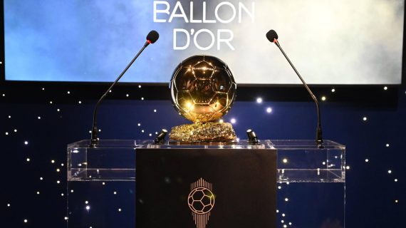 El trofeo dorado del Balón de Oro sobre un atril transparente entre dos micrófonos y un fondo azul con luces blancas