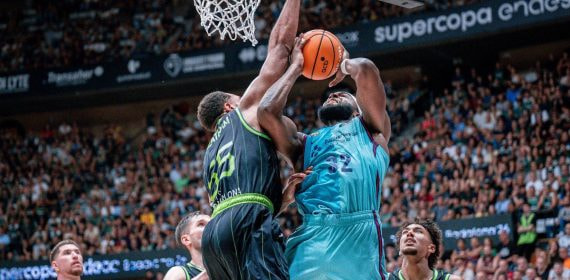 Un jugador del Barça de azul celeste lanza bajo el aro ante el tapón de un jugador del Joventut vestido de negro y verde