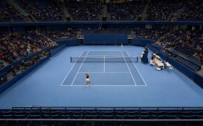 Pista de tenis cubierta azul vista desde la grada alta, con una jugadora a punto de sacar junto a los banquillos