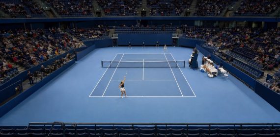 Pista de tenis cubierta azul vista desde la grada alta, con una jugadora a punto de sacar junto a los banquillos
