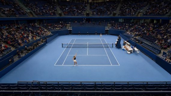 Pista de tenis cubierta azul vista desde la grada alta, con una jugadora a punto de sacar junto a los banquillos