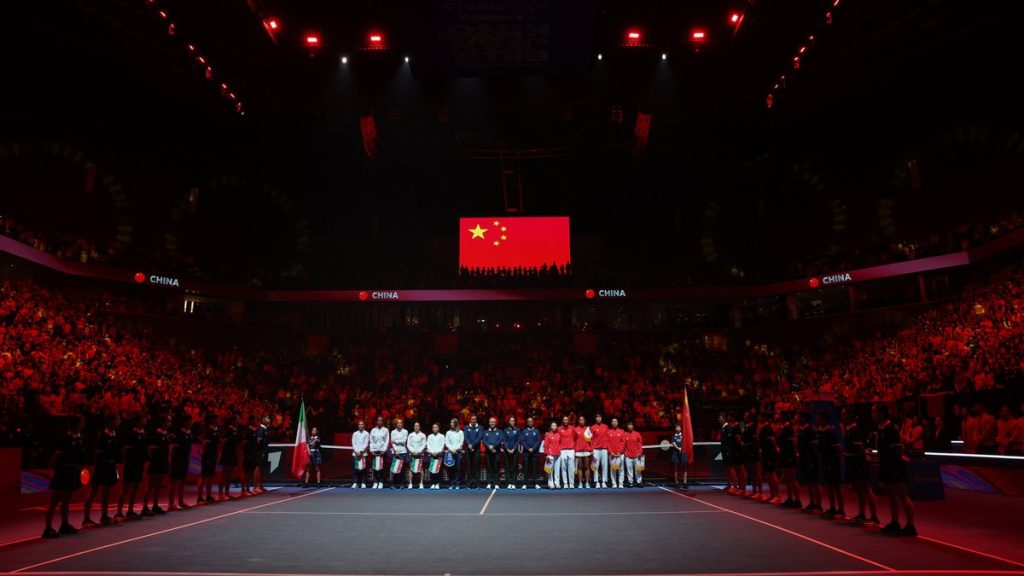 Las selecciones de Italia y China, alineadas en la pista bajo la bandera china durante los himnos en un pabellón en penumbra