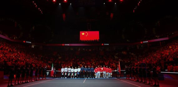 Las selecciones de Italia y China, alineadas en la pista bajo la bandera china durante los himnos en un pabellón en penumbra