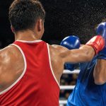 Un boxeador de rojo, de espaldas, conecta un directo a su rival de azul, que se cubre en un ring de competición