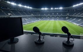 Una cabina de comentaristas con dos micrófonos sobre la mesa y el campo de fútbol iluminado y vacío al fondo