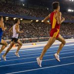 Un mediofondista con equipación roja y amarilla encabeza el grupo en la recta de meta de un estadio de atletismo iluminado