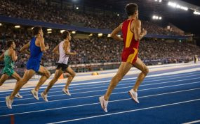 Un mediofondista con equipación roja y amarilla encabeza el grupo en la recta de meta de un estadio de atletismo iluminado