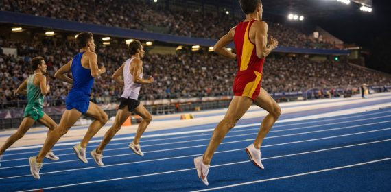 Un mediofondista con equipación roja y amarilla encabeza el grupo en la recta de meta de un estadio de atletismo iluminado