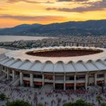 Estadio mediterráneo con cubierta blanca en forma de concha al atardecer, con aficionados camino de la entrada