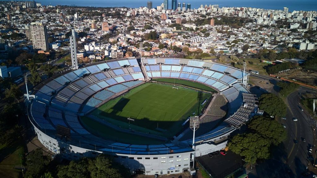 Vista aérea del Estadio Centenario, con sus gradas celestes, rodeado de edificios de Montevideo y el Río de la Plata al fondo