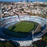 Vista aérea del Estadio Centenario, con sus gradas celestes, rodeado de edificios de Montevideo y el Río de la Plata al fondo
