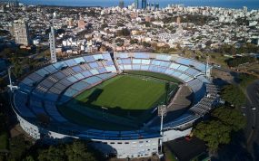 Vista aérea del Estadio Centenario, con sus gradas celestes, rodeado de edificios de Montevideo y el Río de la Plata al fondo