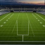 Campo de fútbol americano vacío con las líneas de yardas marcadas y los focos encendidos al anochecer