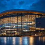 Exterior de un pabellón deportivo moderno iluminado al anochecer con la ciudad al fondo