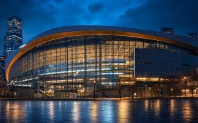 Exterior de un pabellón deportivo moderno iluminado al anochecer con la ciudad al fondo