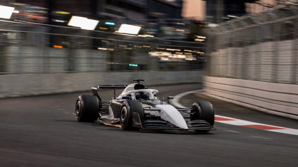 Un monoplaza de Fórmula 1 gris y blanco, sin patrocinadores, traza una curva de un circuito urbano al atardecer