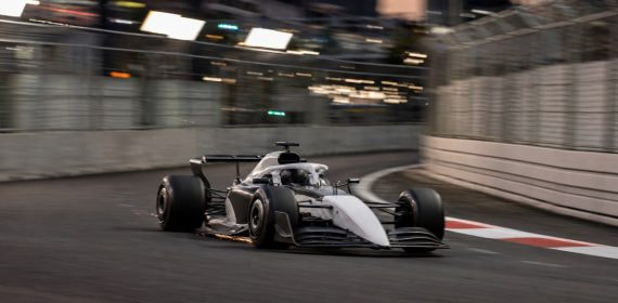 Un monoplaza de Fórmula 1 gris y blanco, sin patrocinadores, traza una curva de un circuito urbano al atardecer