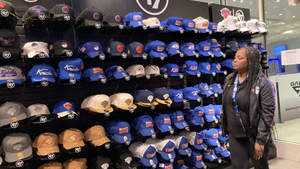 Una dependienta observa una pared entera de gorras de los New York Knicks en tonos azules y naranjas dentro de una tienda