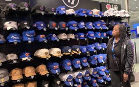 Una dependienta observa una pared entera de gorras de los New York Knicks en tonos azules y naranjas dentro de una tienda