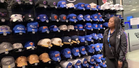 Una dependienta observa una pared entera de gorras de los New York Knicks en tonos azules y naranjas dentro de una tienda