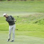 Un golfista con gorra blanca y jersey oscuro termina un golpe de hierro en una calle de hierba verde con un talud al fondo