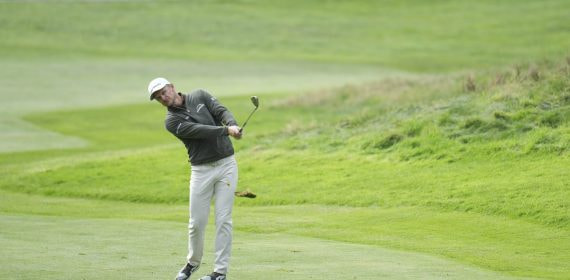 Un golfista con gorra blanca y jersey oscuro termina un golpe de hierro en una calle de hierba verde con un talud al fondo