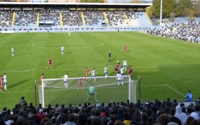 Dos futbolistas saltan a por un balón dividido en el área durante un partido en un estadio con la grada a medio llenar